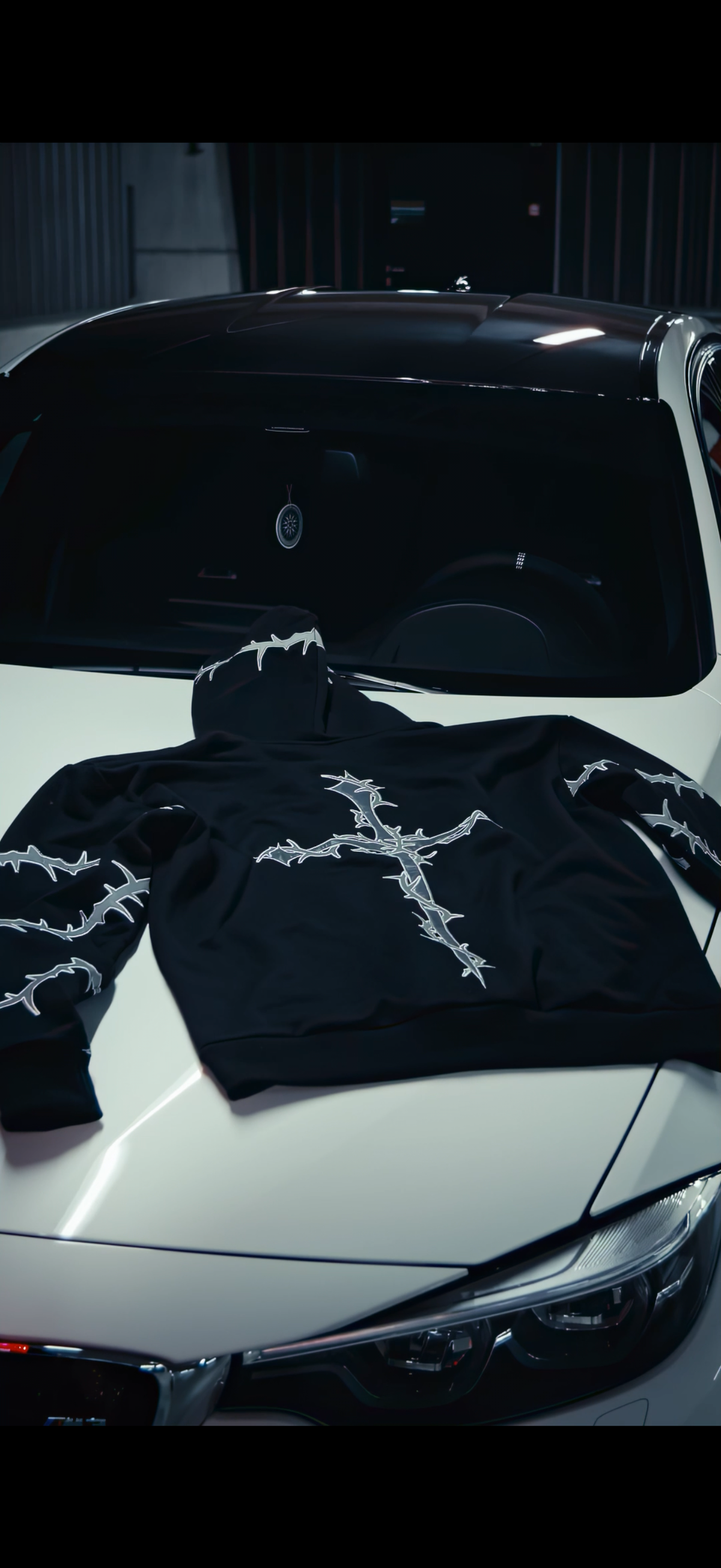 A black hoodie with a white barbed-wire cross graphic on the chest and sleeve details, laid flat on the hood of a white car.