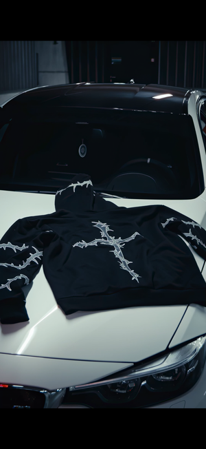 A black hoodie with a white barbed-wire cross graphic on the chest and sleeve details, laid flat on the hood of a white car.