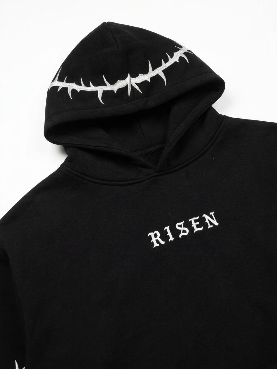 A close-up of a black hoodie featuring a white thorny crown graphic on the hood and the word 'RISEN' in white gothic lettering on the chest, set against a plain white background.